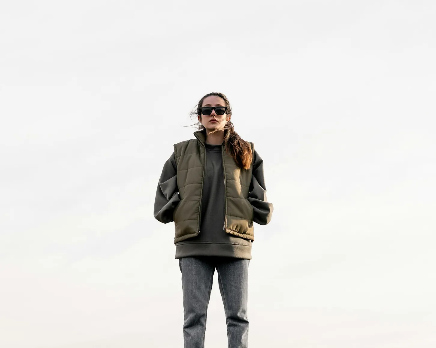 Woman in an olive quilted vest and sunglasses against a pale sky
